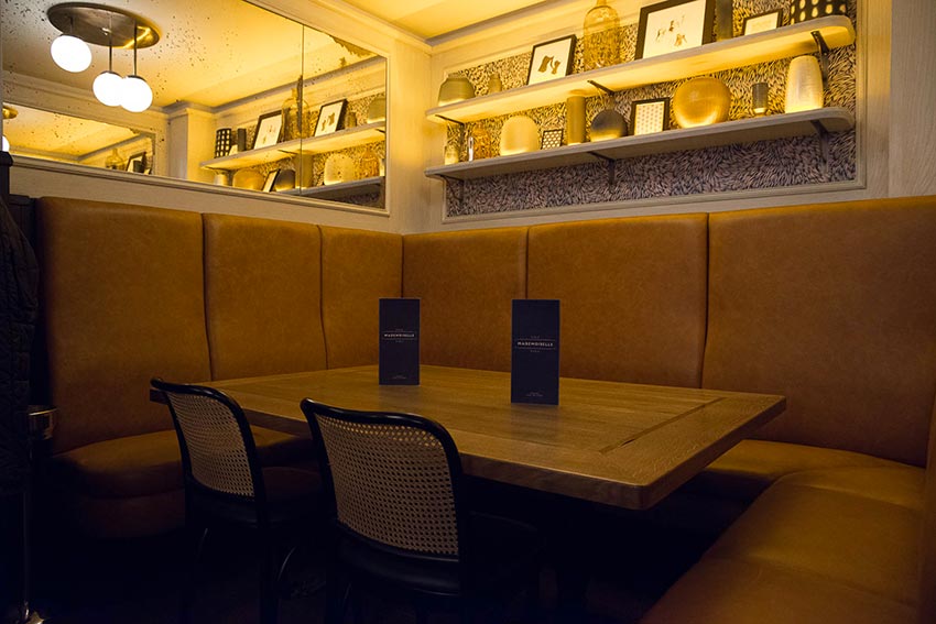 Banquette au dossier haut en cuir naturel et table d'hôtes du Café Mademoiselle de Paris avec son étagère et papier peint Petite Friture.